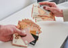 Ein Mann (l) und eine Frau halten unterschiedlich viele Geldscheine und Karten in der Hand.